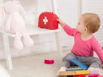Toddler reaching for red first aid kit toy beside rabbit plushie on chair in nursery room.