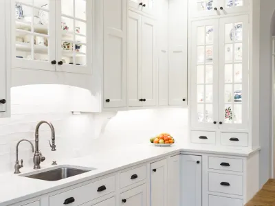 A close-up of a bright kitchen featuring white inset cabinetry with a glass-front display cabinet showcasing decorative dishware.