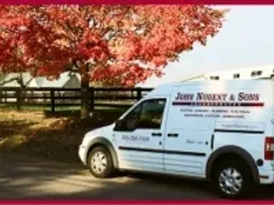 A white John Nugent & Sons service van is parked beside a fence and red-leaved trees on a sunny day. Business signage and contact details are visible on the van.