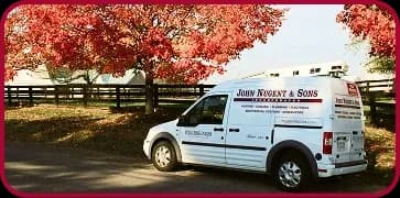 A white John Nugent & Sons service van is parked beside a fence and red-leaved trees on a sunny day. Business signage and contact details are visible on the van.