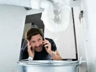 Worried Man Calling Plumber While Watching Water Leaking From Sink