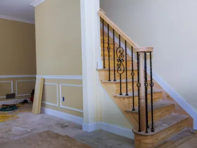Staircase with wooden steps and decorative black iron railing in a partially finished room, showcasing custom cabinetry and trim work by Ace Kustoms.