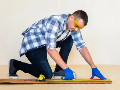 Man measuring wood with a tape measure and square, wearing safety goggles and gloves, illustrating craftsmanship in custom trim work.