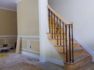 Staircase with wooden steps and decorative black iron railing in a partially finished room, showcasing custom cabinetry and trim work by Ace Kustoms.