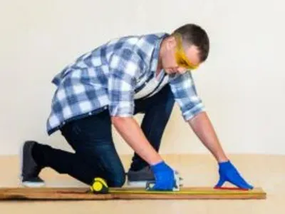 Man measuring wood with a tape measure and square, wearing safety goggles and gloves, illustrating craftsmanship in custom trim work.