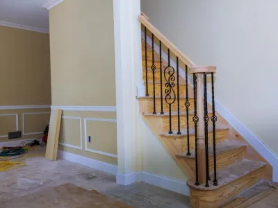 Staircase with wooden steps and decorative black iron railing in a partially finished room, showcasing custom cabinetry and trim work by Ace Kustoms.