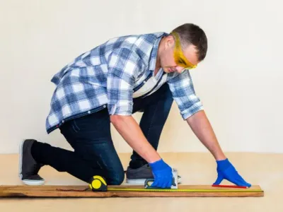 Man measuring wood with a tape measure and square, wearing safety goggles and gloves, illustrating craftsmanship in custom trim work.