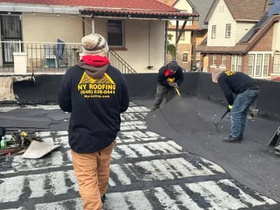 Three workers wearing NY Roofing jackets repair a flat rooftop, using tools to remove old roofing material. Houses with pitched roofs are visible in the background.