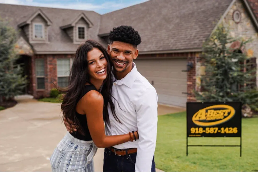 Couple smiling in front of a residential home with A-Best Roofing sign, showcasing roofing services in Tulsa, OK.