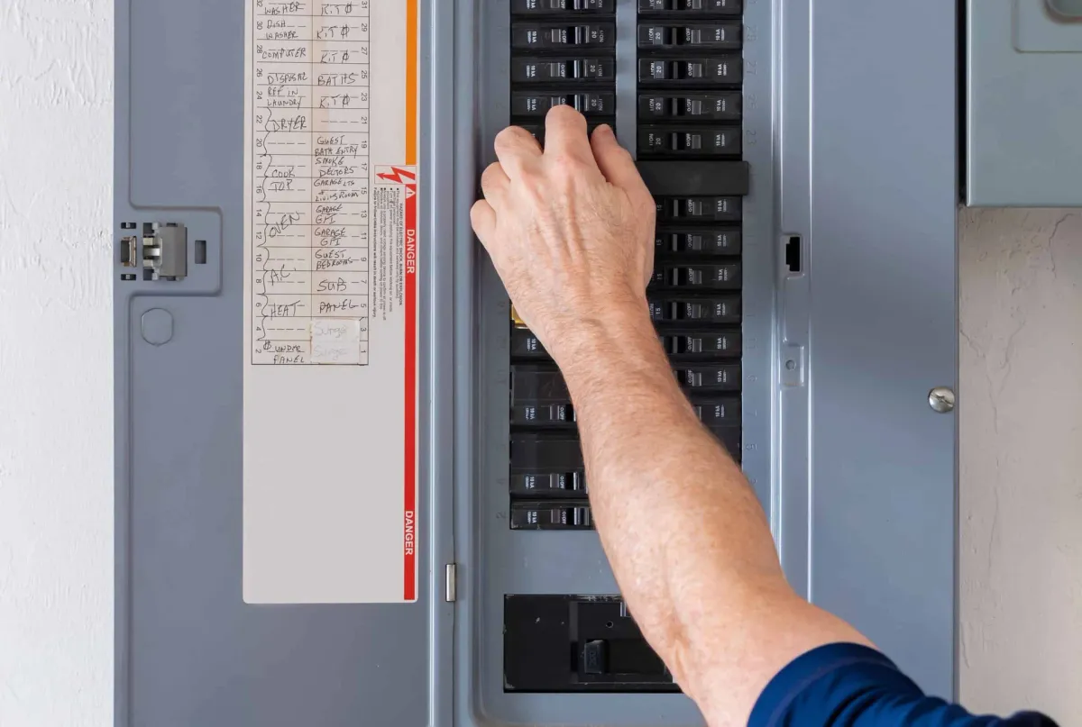 A person’s hand is adjusting a switch inside an electrical breaker panel, with a list of circuit labels visible on the panel door.