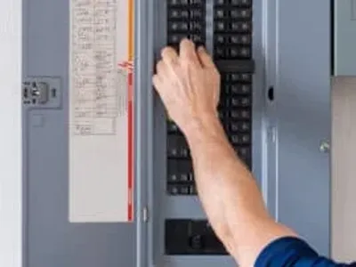 A person’s hand is adjusting a switch inside an electrical breaker panel, with a list of circuit labels visible on the panel door.
