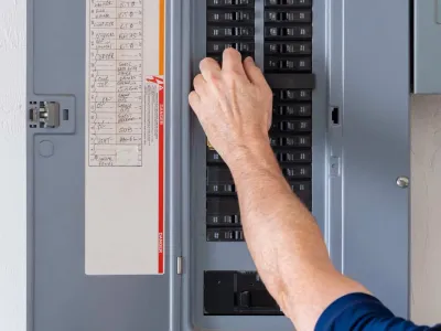 A person’s hand is adjusting a switch inside an electrical breaker panel, with a list of circuit labels visible on the panel door.