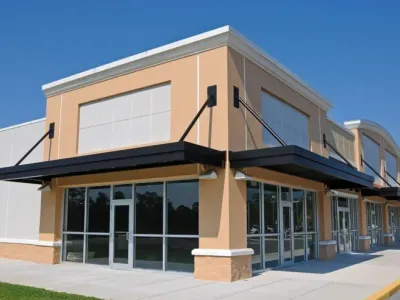 Corner view of a newly built commercial strip mall with tan stucco exterior, black metal awnings, and glass storefronts under a clear blue sky.