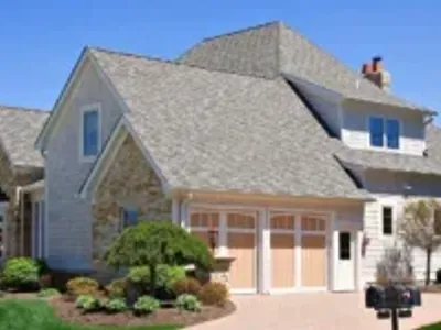 Suburban home exterior with gray asphalt shingle roof, stone and shake siding, three-car garage, and manicured landscaping.