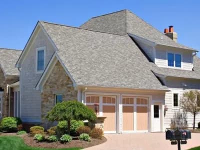Suburban home exterior with gray asphalt shingle roof, stone and shake siding, three-car garage, and manicured landscaping.