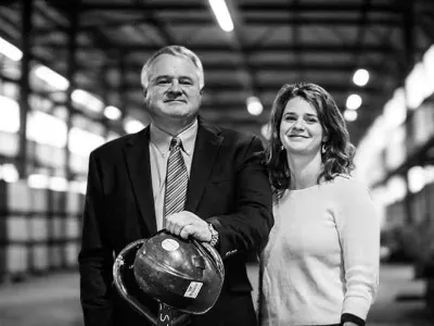 Two business leaders standing in an industrial facility, one holding a hard hat, representing leadership in electrical construction.