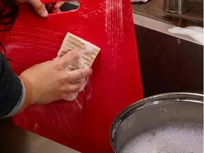 Red cutting board being cleaned in kitchen sink with sponge