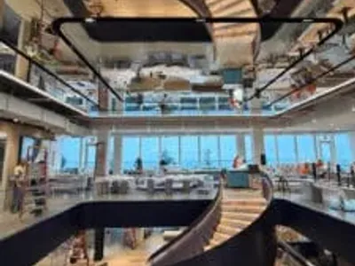 A modern, open office space features a dramatic spiral staircase with black railings and light wood steps. The staircase and workers are reflected in a large mirrored ceiling above.