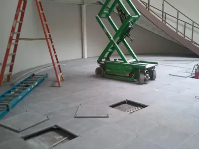 A room under construction with an open raised floor showing cables, a green scissor lift, two ladders (one red, one blue), and a staircase in the background. Some floor panels are removed.