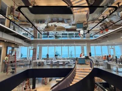 A modern, open office space features a dramatic spiral staircase with black railings and light wood steps. The staircase and workers are reflected in a large mirrored ceiling above.