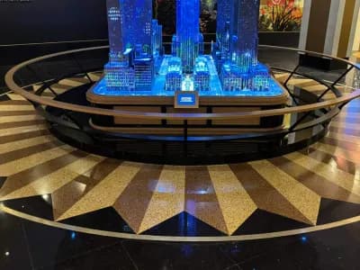 A detailed miniature model of a city skyline, illuminated with blue lights, is displayed on a circular platform with a shining starburst pattern on the black and gold floor.