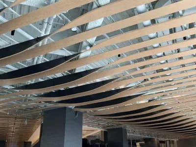 Curved wooden slats form a wave-like pattern on the ceiling of a modern, spacious interior with exposed ductwork above and tables and chairs below.