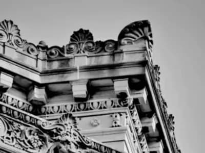 Black and white photo of an ornate building corner, showcasing detailed carvings of faces, floral patterns, and decorative stonework against a clear sky.