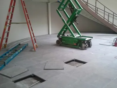 A room under construction with an open raised floor showing cables, a green scissor lift, two ladders (one red, one blue), and a staircase in the background. Some floor panels are removed.