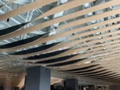 Curved wooden slats form a wave-like pattern on the ceiling of a modern, spacious interior with exposed ductwork above and tables and chairs below.