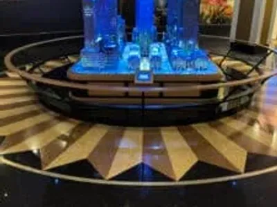 A detailed miniature model of a city skyline, illuminated with blue lights, is displayed on a circular platform with a shining starburst pattern on the black and gold floor.