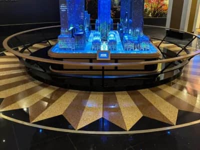 A detailed miniature model of a city skyline, illuminated with blue lights, is displayed on a circular platform with a shining starburst pattern on the black and gold floor.