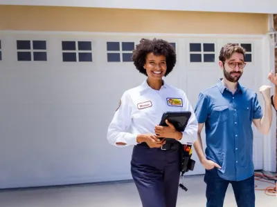 Female electrician with a tablet smiling at the camera next to two customers in front of a garage.